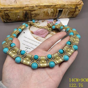 May include: A gold-toned statement necklace featuring round turquoise-colored beads with dark veining, interspersed with textured metal segments and small clear crystals. The necklace includes a chain extension and measures 14 cm plus 9 cm.