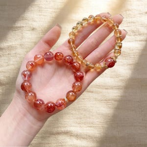 May include: Two beaded bracelets. One bracelet features reddish-orange beads, while the other has yellow-orange beads. The bracelets are displayed on a light-colored surface. The beads appear to be semi-transparent.
