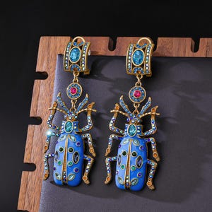 May include: A pair of drop earrings featuring a beetle design in bright blue enamel with gold-tone metal accents. Each beetle is embellished with small multicolored crystals and a central oval blue stone, suspended from a rectangular top piece.