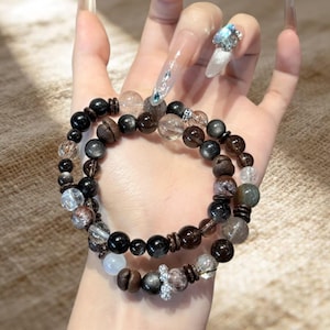 Puede incluir: Se muestran dos pulseras de cuentas con una variedad de cuentas oscuras, incluyendo negras, marrones y transparentes. Las pulseras están superpuestas, mostrando las diferentes texturas y tamaños de las cuentas. Un pequeño dije decorativo es visible en una pulsera.