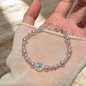 May include: A bracelet featuring a series of lustrous white pearls interspersed with small silver-toned beads. The design includes two small, circular, crystal-encrusted spacers and a central square-shaped charm with a clear, faceted stone.