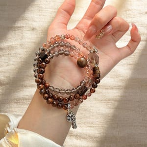 May include: A set of three beaded bracelets in shades of brown, clear, and pink. The bracelets feature various beads, including round, faceted, and wooden beads. One bracelet has a charm with Chinese characters.