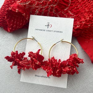 May include: Gold hoop earrings featuring bright red floral detailing. The earrings are presented on a card with the text "JAPANESE CRAFT STORIES" and the handle "@wacraftstories". A red fabric with a white pattern is in the background.