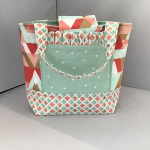 May include: A handmade tote bag with a geometric pattern in coral, mint green, and tan. The bag features a mint green pocket with white triangles and matching handles. The bottom has a diamond pattern.