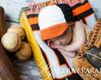Può includere: Un neonato con un berretto da baseball all'uncinetto nero, bianco e arancione, appoggiato su una coperta a tema baseball. Un guanto da baseball, palline da baseball e una mazza di legno sono nelle vicinanze. Fotografia professionale.