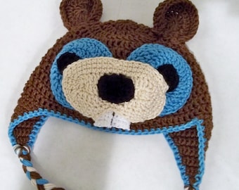 May include: A brown crochet hat with large, round ears and a cartoon-like face. The face features light blue eyes, a tan nose, black nostrils, and white teeth. The hat has a braided chin strap with blue, brown, and white strands.