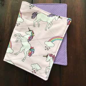 May include: Two quilted fabric coasters. The top coaster is light pink with a unicorn and rainbow pattern. The bottom coaster is purple with white polka dots. The coasters are square with rounded corners.