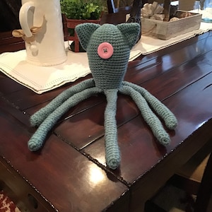 Puede incluir: Una criatura de ganchillo teal hecha a mano con un ojo de botón rosa y múltiples tentáculos. El juguete de peluche caprichoso tiene dos orejas pequeñas y se exhibe sobre una mesa de madera oscura. El juguete es un artículo único y artesanal.