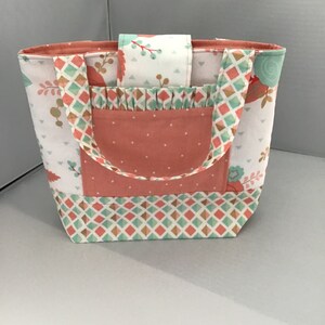 May include: A handmade tote bag with a floral and geometric pattern. The bag features a peach-colored pocket and handles, with a ruffled accent. The main fabric is white with a pattern of peach, teal, and gold.