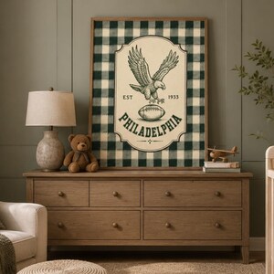 Pu&ograve; includere: Un quadro rettangolare incorniciato con uno sfondo a quadri scozzesi verde scuro e bianco. Al centro, un emblema color crema presenta un'illustrazione dettagliata di un'aquila, un pallone da football e il testo EST 1933 PHILADELPHIA.