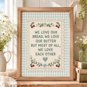 May include: A framed artwork with a light brown wooden frame. The artwork features a light blue and white gingham background with a central embroidered design. The design includes text that reads "WE LOVE OUR BREAD, WE LOVE OUR BUTTER BUT MOST OF ALL, WE LOVE EACH OTHER."