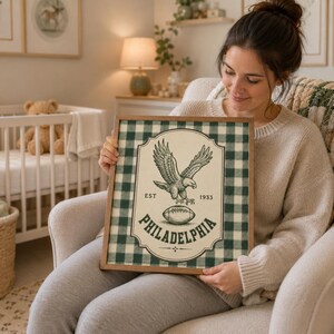 Pu&ograve; includere: Una stampa artistica incorniciata con uno sfondo a quadri verde scuro e bianco. Il design centrale mostra un'illustrazione dettagliata di un'aquila sopra un pallone da football, con il testo EST 1933 e PHILADELPHIA stampato in inchiostro verde.