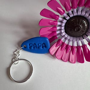 May include: A blue keychain with the word "PAPA" embossed on it, attached to a silver keyring. A large, decorative flower with pink and purple petals is in the background. The flower has a dark brown center.