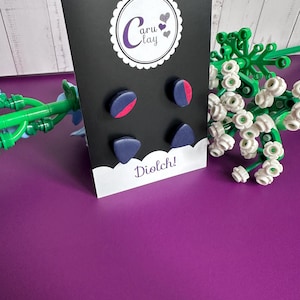 May include: A black card displays four pairs of handmade clay earrings in navy blue and red. The card has a white scalloped border with the text "Caru Clay" and "Diolch!" on the bottom. The background is a solid purple.
