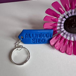 May include: A blue keychain with the text "ALLWEOOI SIED" attached to a silver keyring. A pink and purple decorative object is in the background.