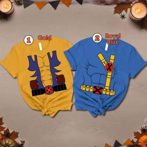 May include: Two short-sleeved t-shirts featuring superhero costume graphics. The gold shirt displays a blue vest, brown gloves, and a black belt with a red X buckle. The royal blue shirt features a yellow harness, a red X emblem, and a yellow utility belt.