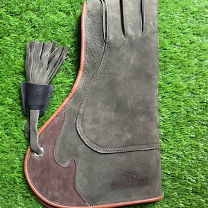 Op de afbeelding: Een grijze leren valkeniershandschoen met een bruin accent en oranje bies. De handschoen heeft een vingerstalontwerp en een leren kwastje. De tekst "DREAM BIRD" is zichtbaar op de handschoen.