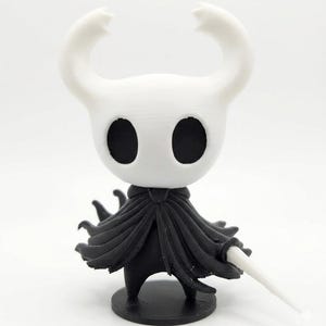 Puede incluir: Figura impresa en 3D de un personaje con cabeza blanca y grandes ojos negros, que lleva una capa negra y sostiene una espada blanca. La figura tiene dos cuernos curvados y se apoya sobre una base negra.