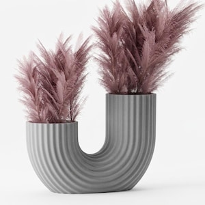 May include: A modern, gray, U-shaped vase with a ribbed texture, holding two bunches of fluffy, mauve-colored pampas grass. The vase has a unique, curved design, and the background is plain white.