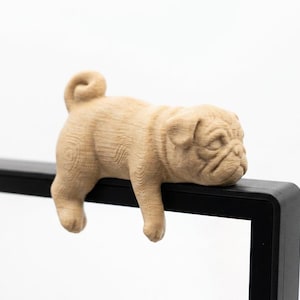 May include: A light brown, wooden pug dog figurine, carved with detailed fur texture, is resting on a black surface. The pug's head and front paws are hanging over the edge, giving it a relaxed appearance. The background is plain white.
