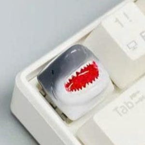 May include: A custom-made shark keycap with a gray top, white body, and a red, toothy mouth. The keycap is installed on a white keyboard, next to other white keys.