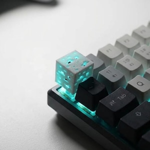 Peut inclure: Gros plan sur un clavier mécanique avec une touche personnalisée en forme de cube blanc avec un intérieur bleu lumineux. Le clavier a des touches noires et grises.