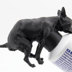 Könnte beinhalten: Eine schwarze Hundefigur, die auf einer Zahnpastatube sitzt. Der Hund ist in einer realistischen Pose modelliert, mit den Vorderpfoten auf der Tube. Die Zahnpastatube ist weiß mit blauen Akzenten und Text.