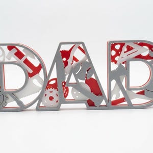 Pode incluir: Uma placa decorativa que soletra "DAD" em letras cinzentas com detalhes em vermelho e branco. As letras são preenchidas com várias formas de ferramentas e engrenagens, criando um design único. Um presente perfeito para o Dia dos Pais.