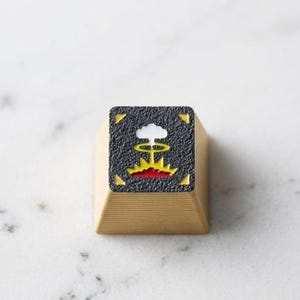 May include: A beige keycap with a textured black square featuring a colorful graphic of a mushroom cloud, sun, and red base. The keycap has yellow corner accents and is likely for a mechanical keyboard.