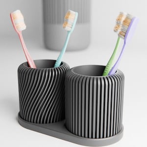 May include: Two grey toothbrush holders with toothbrushes. One has a spiral design, the other has vertical lines. The toothbrushes are pink, blue, and green with white bristles. A third, similar grey container is in the background.