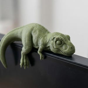 May include: A green Tyrannosaurus Rex figurine, resting on a black surface. The dinosaur has detailed skin texture and closed eyes, giving it a relaxed appearance. The tail curves down, adding to the figurine's playful design.