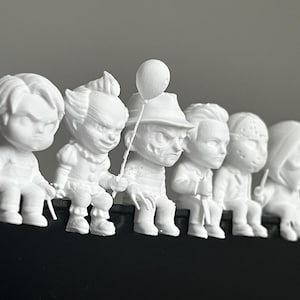 May include: A row of white figurines depicting horror movie characters, including Chucky, Pennywise, Freddy Krueger, and others. Each figure is seated, with detailed facial expressions and iconic features. The figurines are arranged on a black surface.