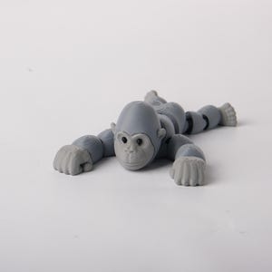 Op de afbeelding: Een grijs gearticuleerd gorilla-speelgoed, liggend op een wit oppervlak. Het speelgoed heeft een gedetailleerd gezicht, gearticuleerde ledematen en een gesegmenteerd lichaam. De handen en voeten van de gorilla zijn in de vorm van een vuist.