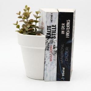 Puede incluir: Una maceta de cerámica blanca con una pequeña planta verde, utilizada como sujetalibros. Dos libros están de pie junto a la maceta. Un libro tiene el título "KALTE STILLE" y el otro "IN DER FINSTERNIS".