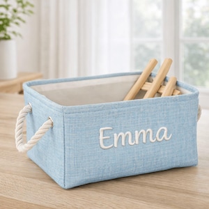 May include: A light blue rectangular storage bin with the name "Emma" embroidered in white. The bin has rope handles and contains wooden items. The bin is sitting on a wooden surface.