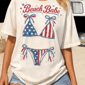 May include: Cream-colored t-shirt with a patriotic bikini graphic. The bikini design features red and white stripes and blue with white stars. The text "Beach Babe" is above the bikini graphic.