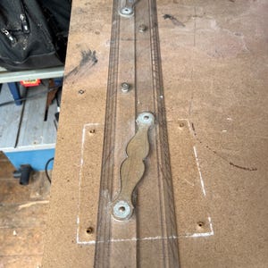 May include: A long, clear, rectangular drafting ruler featuring two attached metallic handles with a decorative, wavy edge. The ruler is marked with precise measurement lines along its edges and is secured to a flat surface with small screws.