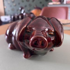 May include: A glossy, dark brown ceramic figurine of a pig. The pig is depicted in a standing position with its eyes closed and ears drooping forward. The surface of the ceramic has a smooth, reflective finish that highlights the rounded contours.