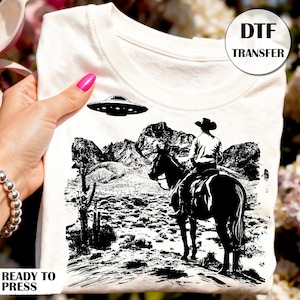 May include: White t-shirt featuring a black and white graphic of a cowboy on horseback with a UFO above a desert landscape. The shirt has the text "READY TO PRESS" at the bottom and "DTF TRANSFER" in a circle at the top right.