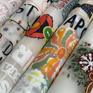 May include: A collection of rolled-up patterned papers, featuring various designs. Some rolls display text like "STARS" and "WHITE CHRISTMAS." Other designs include a retro flower, a cartoon character, and holiday-themed graphics. The papers are in shades of white, beige, and various colors.
