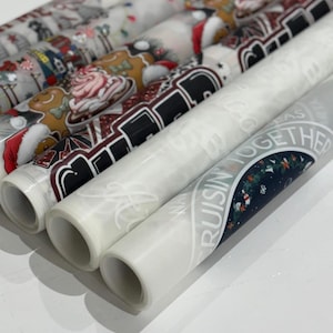 May include: Several rolls of decorative wrapping paper with holiday-themed designs. The patterns include Santa Claus, gingerbread men, and festive text. The rolls are white and have various colors.