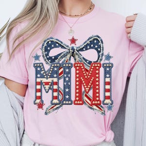 May include: Pink t-shirt featuring a patriotic design with the word "MIMI" in red, white, and blue. The design includes a bow and stars, creating a festive and celebratory look. The shirt is a casual wear item.