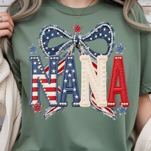 May include: Sage green t-shirt featuring a patriotic design. The design includes the word "NANA" in red, white, and blue, with a bow and stars. The letters are embellished with pearls and the bow has a denim-like texture.