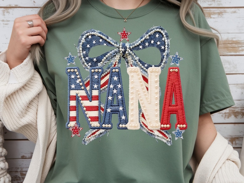 May include: Sage green t-shirt featuring a patriotic design. The design includes the word "NANA" in red, white, and blue, with a bow and stars. The letters are embellished with pearls and the bow has a denim-like texture.