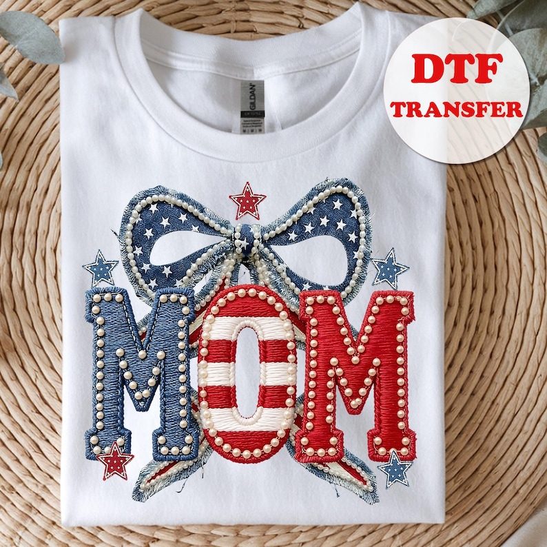 May include: White t-shirt featuring a patriotic design with the word "MOM" in red, white, and blue. The design includes a bow and stars, with pearl accents. The shirt is a DTF transfer.