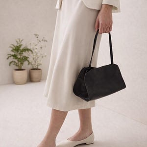 May include: A black handbag with a rectangular shape and curved top is held by a person. The person is wearing a cream-colored skirt, blazer, and ballet flats. Two small potted plants are in the background.