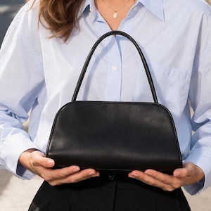 Minimalist black leather shoulder bag with smooth finish and curved baguette handbag shape, women leather bag featuring top handle and detachable strap for crossbody bag use, elegant aesthetic bag inspired by vintage baguette bag and 90s underarm bag trend, sleek structured leather bag perfect for everyday bag use, lightweight leather purse with zipper closure, classic handbag for daily essentials, pinterest bag style with quiet luxury feel, minimalist work bag and trendy everyday bag ideal gift