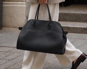 May include: A large black leather handbag with a rounded top silhouette, dual top handles, and side buckle straps. The bag features a textured finish and a spacious main compartment, providing a structured yet functional design for carrying daily essentials.