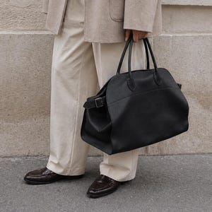 May include: A large black leather handbag with dual top handles and a side buckle strap, paired with a beige wool blazer, a cream ribbed sweater, cream trousers, and dark brown loafers. The handbag features a rounded silhouette and smooth texture.