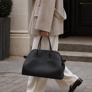 May include: A large black leather handbag with a rounded top silhouette, dual top handles, and side buckle straps. The bag features a textured finish and a spacious main compartment, providing a structured yet functional design for carrying daily essentials.